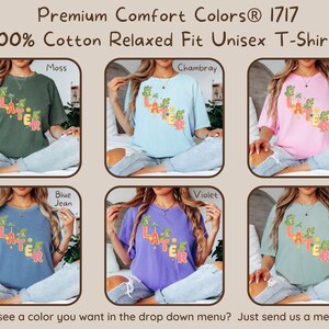 May include: A selection of unisex t-shirts in various colors, including moss green, chambray blue, and blossom pink. Each shirt features the word "LATER" in a colorful, tropical-themed design. The shirts are made of 100% cotton and have a relaxed fit.