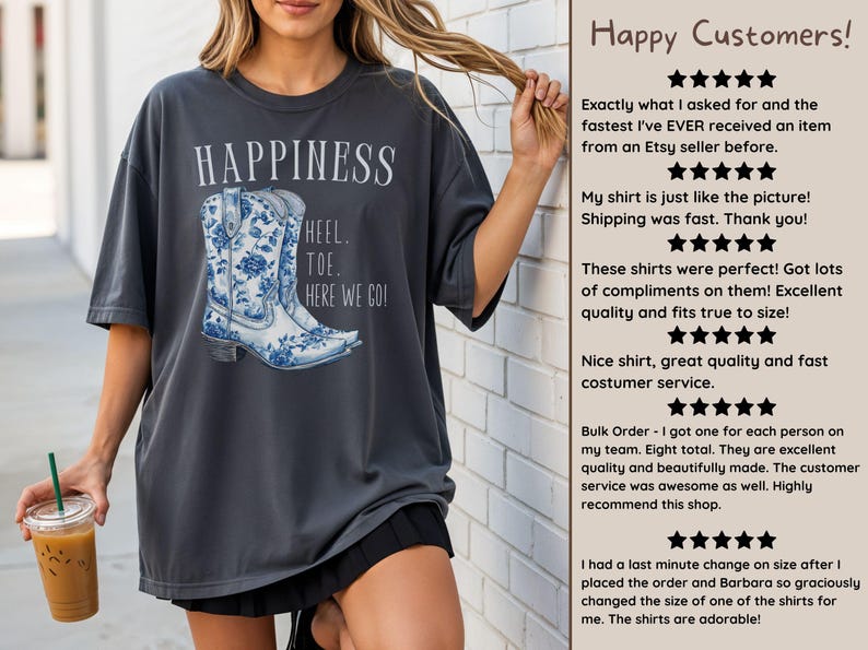 May include: Dark grey t-shirt featuring a blue and white floral cowboy boot graphic with the word "HAPPINESS" above it. The text "HEEL. TOE. HERE WE GO!" is below the boot. The shirt is worn by a person.