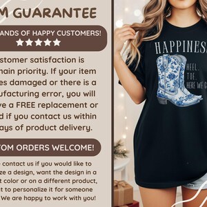 May include: Black t-shirt with the word "HAPPINESS" and a blue and white floral cowboy boot design. The text "HEEL...TOE...HERE WE GO!" is below the boot. A casual wear item.