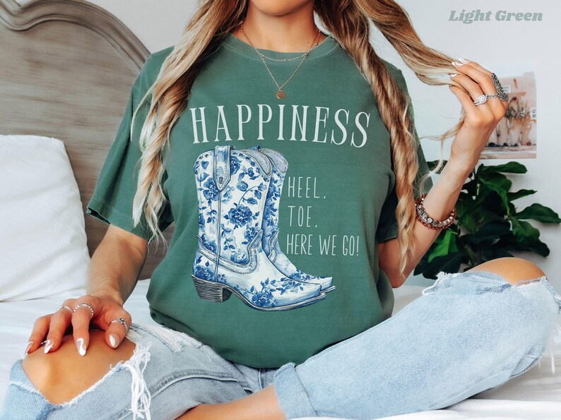 May include: Light green t-shirt featuring the word "HAPPINESS" and a blue and white floral cowboy boot design. The text "HEEL. TOE. HERE WE GO!" is printed below the boots. The shirt is a casual style.