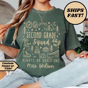 May include: Green t-shirt with a camping-themed design. The shirt features the text "SECOND GRADE Squad" with illustrations of a compass, backpack, mountains, tent, raccoon, and other outdoor elements. The text "ALWAYS AN ADVENTURE Mrs. Wilson" is also printed on the shirt.
