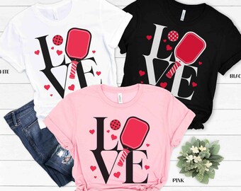 May include: Three t-shirts with the word "LOVE" in black letters with a red and white paddleball and red hearts. The shirts are white, black, and pink.
