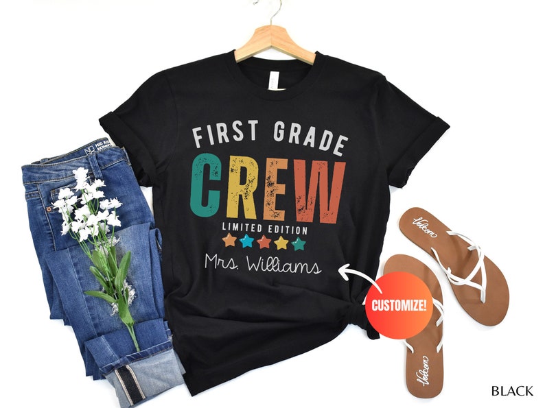 Może przedstawiać: Czarny T-shirt z napisem "First Grade Crew Limited Edition" w retro czcionce z kolorowymi akcentami. Pięć gwiazdek znajduje się pod tekstem, a napis "Mrs. Williams" znajduje się pod gwiazdkami. Napis "CUSTOMIZE!" znajduje się w czerwonym k&oacute;łku.