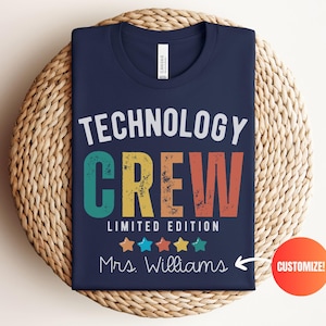 May include: Navy blue t-shirt with the words "TECHNOLOGY CREW" in white, teal, yellow, and orange. The shirt also says "LIMITED EDITION" and "Mrs. Williams" with colorful stars. The shirt is folded on a woven mat.