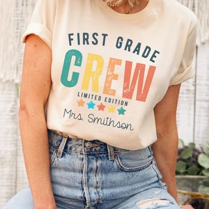 Może przedstawiać: Jasnobeżowa koszulka z napisem "First Grade Crew" w retro kolorystyce tęczy. Pod gł&oacute;wnym tekstem znajduje się napis "Limited Edition", a następnie cztery gwiazdki i napis "Mrs. Smithson".