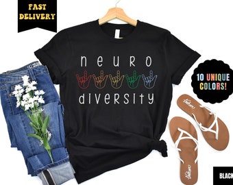 May include: Black t-shirt with the text "neuro diversity" in white. Five colorful hand signs are displayed above the text. The shirt is laid flat on a surface with a pair of brown sandals and blue jeans.