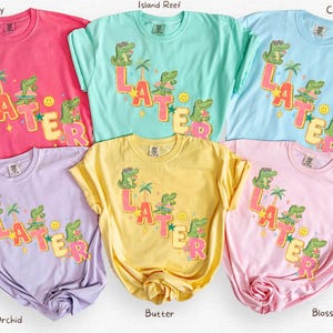 May include: A collection of colorful t-shirts in shades of pink, blue, green, yellow, and purple. Each shirt features a graphic design with the word "LATER" and cartoon alligators, palm trees, and a smiley face. The shirts are casual wear.