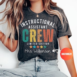 May include: Dark heather grey t-shirt with the words "INSTRUCTIONAL ASSISTANT CREW" in white, teal, yellow, and orange. Below the text are colourful stars and the name "Mrs. Wilkinson". The shirt is a limited edition.