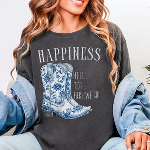 May include: Dark grey t-shirt featuring the word "HAPPINESS" and a graphic of blue and white floral cowboy boots. The text "HEEL. TOE. HERE WE GO!" is below the boots.