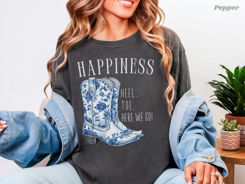 May include: Dark grey t-shirt featuring the word "HAPPINESS" and a graphic of blue and white floral cowboy boots. The text "HEEL. TOE. HERE WE GO!" is below the boots.