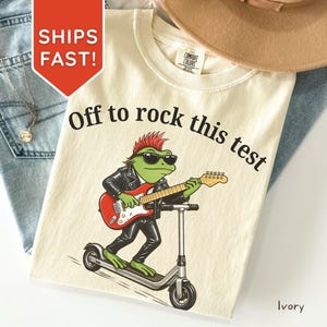 May include: Ivory-colored t-shirt with the text "Off to rock this test." The graphic features a green frog in a leather jacket, sunglasses, and a guitar, riding a scooter. A brown hat and blue jeans are in the background.