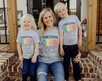 May include: Three people wearing matching grey t-shirts with the words "Homeschool Crew" in colourful letters. The shirts also say "Not all classrooms have four walls." One shirt says "preschool" and another says "kindergarten."