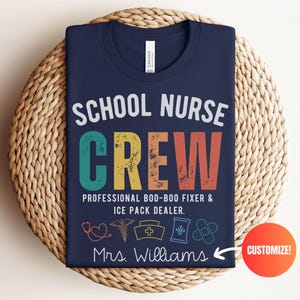 May include: Navy blue t-shirt with the text "SCHOOL NURSE CREW" in white, teal, yellow, and orange. The shirt also says "PROFESSIONAL BOO-BOO FIXER & ICE PACK DEALER" and "Mrs. Williams". A red circle with the word "CUSTOMIZE!" is on the bottom right.