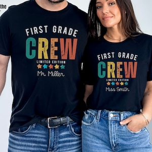 Może przedstawiać: Dwie czarne koszulki z napisem "First Grade Crew" w kolorowych, retro literach. Jedna koszulka ma napis "Mr. Miller", a druga "Miss Smith".