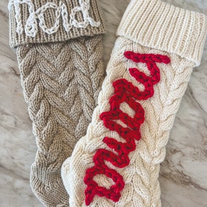 Peut inclure: Deux chaussettes de No&euml;l en tricot avec des motifs en c&acirc;ble. Une chaussette est beige avec le mot "Papa" brod&eacute; en blanc. L'autre chaussette est blanche avec le mot "Maman" brod&eacute; en rouge.