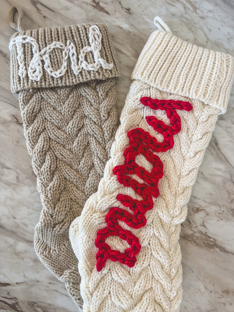 Peut inclure: Deux chaussettes de No&euml;l en tricot avec des motifs en c&acirc;ble. Une chaussette est beige avec le mot "Papa" brod&eacute; en blanc. L'autre chaussette est blanche avec le mot "Maman" brod&eacute; en rouge.