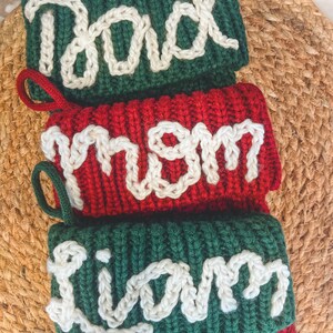 Peut inclure: Trois chaussettes de No&euml;l en tricot avec les noms "Dad", "Mom" et "Liam" brod&eacute;s en fil blanc sur un fond vert. La chaussette pour "Mom" est rouge.