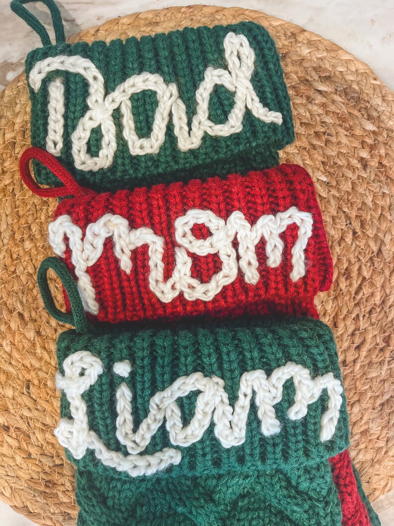 Peut inclure: Trois chaussettes de No&euml;l en tricot avec les noms "Dad", "Mom" et "Liam" brod&eacute;s en fil blanc sur un fond vert. La chaussette pour "Mom" est rouge.