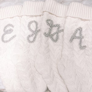 Peut inclure: Quatre chaussettes blanches en tricot avec un motif en c&acirc;ble et des lettres brod&eacute;es grises E, G, J et A.