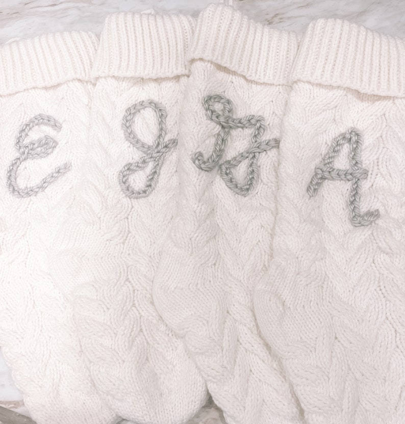 Peut inclure: Quatre chaussettes blanches en tricot avec un motif en c&acirc;ble et des lettres brod&eacute;es grises E, G, J et A.