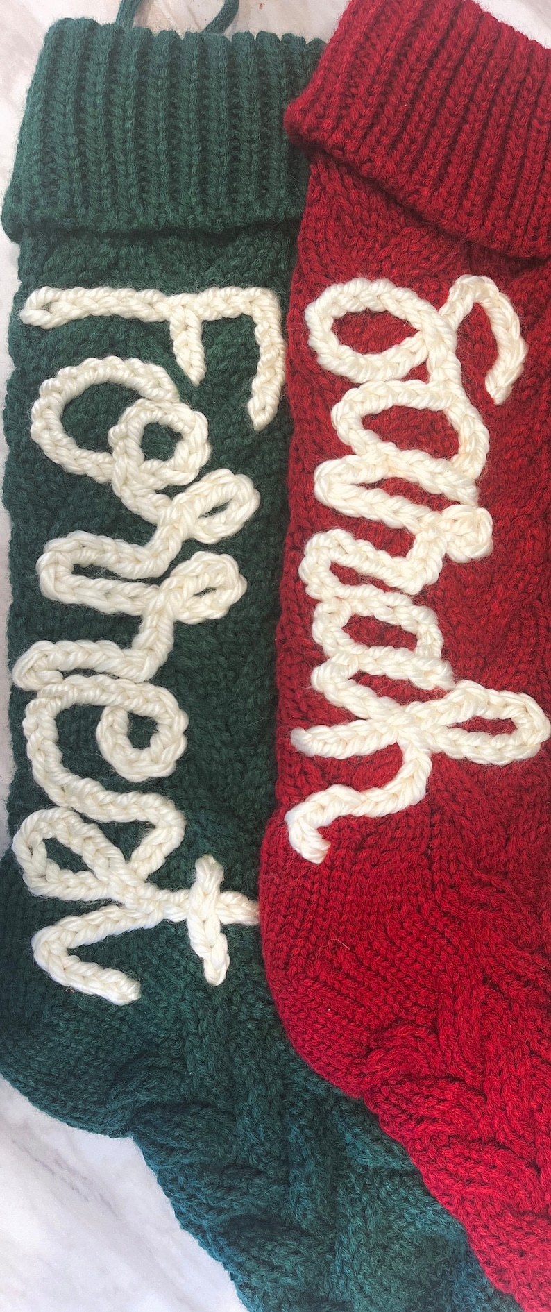 Peut inclure: Deux chaussettes de No&euml;l en tricot avec des lettres en fil blanc. La chaussette de gauche est verte avec le mot "Happy" brod&eacute; dessus. La chaussette de droite est rouge avec le mot "Merry" brod&eacute; dessus.