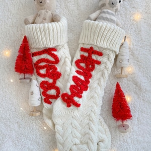 Peut inclure: Deux chaussettes de No&euml;l en tricot blanc avec des lettres brod&eacute;es rouges. Une chaussette a le mot "Merry" brod&eacute; dessus et l'autre a le mot "Happy" brod&eacute; dessus. Chaque chaussette contient un animal en peluche. Une chaussette contient un z&egrave;bre en peluche et l'autre un  ch&egrave;vre en peluche.