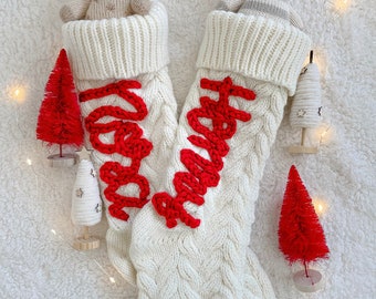 May include: Two white knitted Christmas stockings with red embroidered lettering. One stocking has the word "Merry" embroidered on it and the other has the word "Happy" embroidered on it. Each stocking has a stuffed animal inside. One stocking has a stuffed zebra and the other has a stuffed goat.