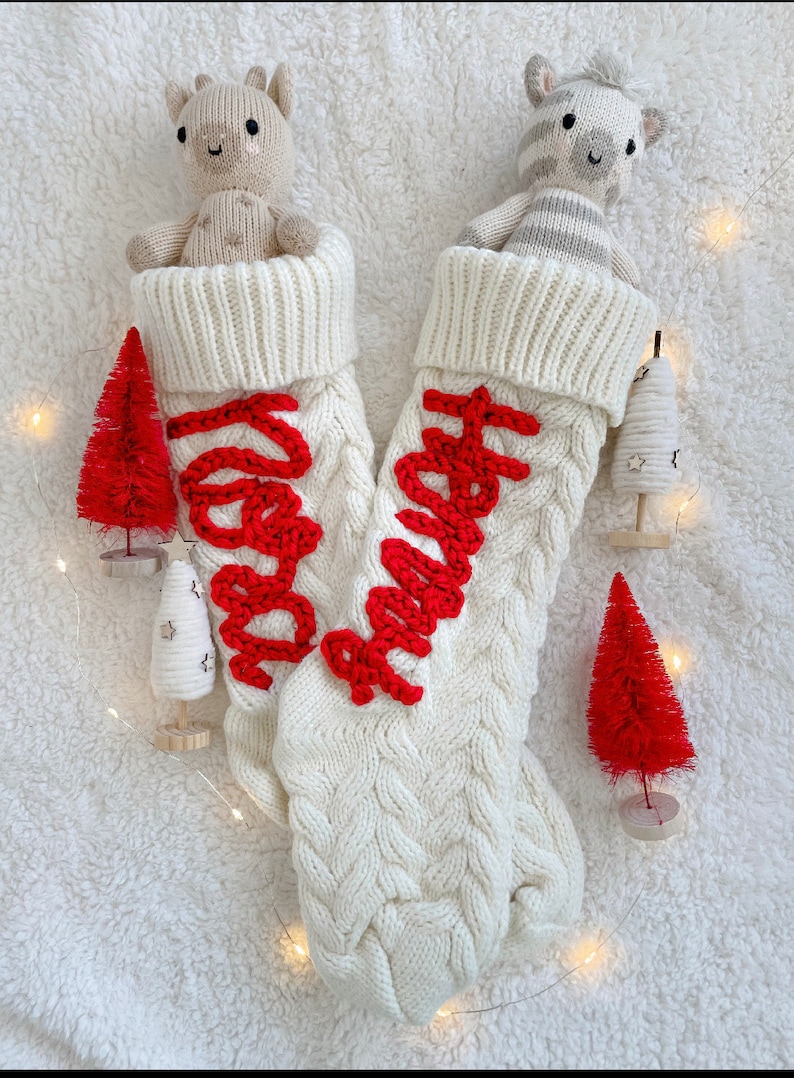 Peut inclure: Deux chaussettes de No&euml;l en tricot blanc avec des lettres brod&eacute;es rouges. Une chaussette a le mot "Merry" brod&eacute; dessus et l'autre a le mot "Happy" brod&eacute; dessus. Chaque chaussette contient un animal en peluche. Une chaussette contient un z&egrave;bre en peluche et l'autre un  ch&egrave;vre en peluche.