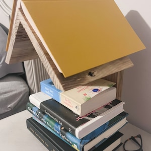 May include: A handmade wooden book shelf shaped like a house, with a yellow book as the roof. Several books are stacked on the shelf, including titles like "Hello Summer" and "Little Fires Everywhere". Tortoiseshell glasses rest on the table.