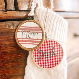 May include: Two embroidered hoop ornaments with wooden frames. One features red and white striped fabric with the word "Mom" stitched in green thread. The other has red and white gingham fabric with "Dad" stitched in green.