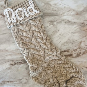Peut inclure: Une chaussette de No&euml;l en tricot beige avec un motif en c&acirc;ble blanc. La chaussette porte le nom "Papa" en feutre blanc sur le revers.