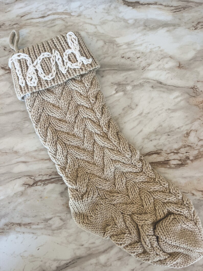 Peut inclure: Une chaussette de No&euml;l en tricot beige avec un motif en c&acirc;ble blanc. La chaussette porte le nom "Papa" en feutre blanc sur le revers.