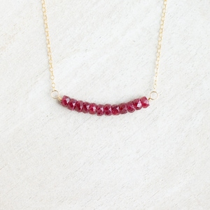 May include: A delicate gold chain necklace featuring a bar of faceted, deep red gemstones. The gemstones are arranged in a curved line, suspended from the gold chain. The necklace is displayed on a white surface.
