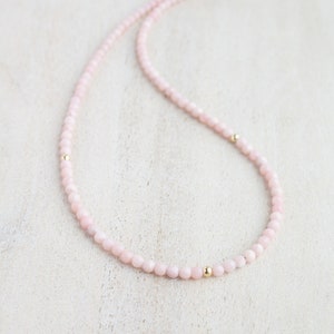 May include: A delicate necklace featuring small, round, pale pink beads. The necklace is accented with small, gold-colored beads and a gold-colored clasp. The jewelry is displayed on a light-colored surface.