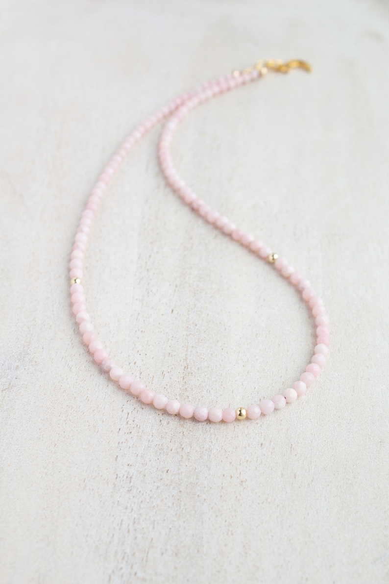 May include: A delicate necklace featuring small, round, pale pink beads. The necklace is accented with small, gold-colored beads and a gold-colored clasp. The jewelry is displayed on a light-colored surface.