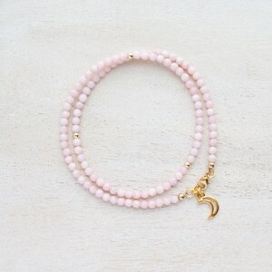 May include: A delicate necklace featuring small, light pink beads. The necklace has gold-colored accents and a gold crescent moon charm. The necklace is displayed on a white, textured surface.