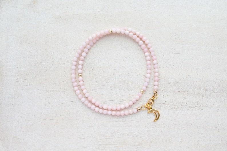 May include: A delicate necklace featuring small, light pink beads. The necklace has gold-colored accents and a gold crescent moon charm. The necklace is displayed on a white, textured surface.