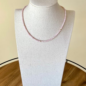 May include: A delicate necklace featuring small, light pink beads strung together. The necklace has gold-colored accents and is displayed on a white jewelry bust. The necklace is a simple, elegant piece of jewelry.