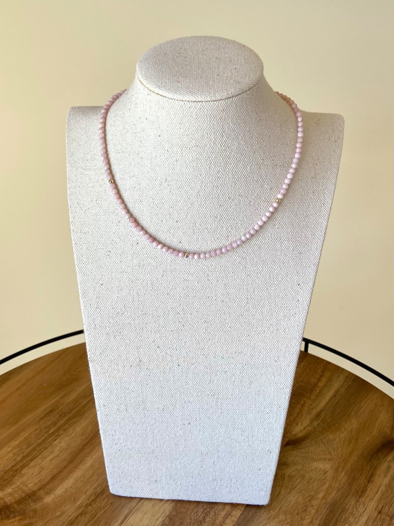 May include: A delicate necklace featuring small, light pink beads strung together. The necklace has gold-colored accents and is displayed on a white jewelry bust. The necklace is a simple, elegant piece of jewelry.