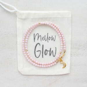 May include: A delicate pink beaded necklace with gold accents and a crescent moon charm, displayed on a small canvas bag. The bag has the words "Mellow Glow" printed on it.