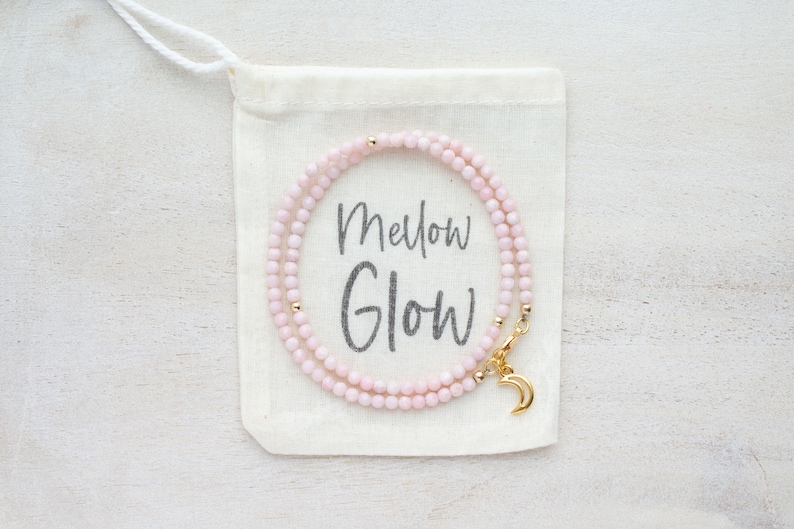 May include: A delicate pink beaded necklace with gold accents and a crescent moon charm, displayed on a small canvas bag. The bag has the words "Mellow Glow" printed on it.