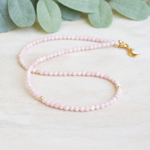 May include: Delicate pink beaded necklace with gold accents. The necklace features small, round pink beads strung together, with gold beads interspersed. A gold-colored crescent moon charm hangs from the clasp.