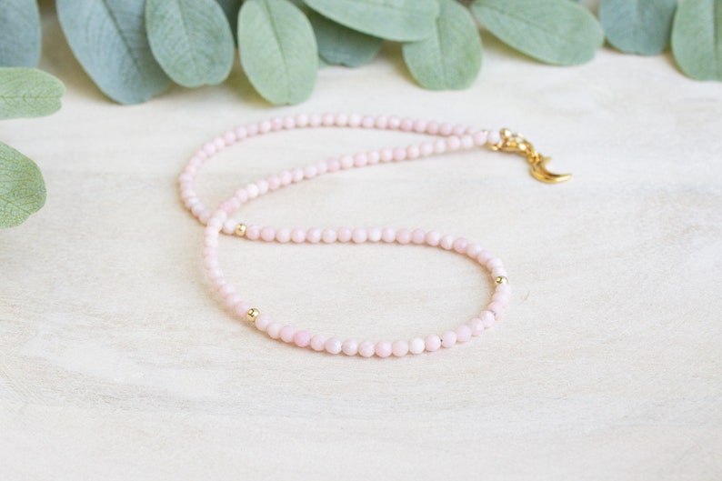 May include: Delicate pink beaded necklace with gold accents. The necklace features small, round pink beads strung together, with gold beads interspersed. A gold-colored crescent moon charm hangs from the clasp.
