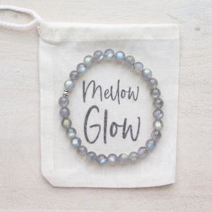 Puede incluir: Un brazalete de piedra de labradorita gris e iridiscente con un acento de cuentas plateadas. El brazalete está en una bolsa de cordón blanco con el texto "Mellow Glow" impreso en ella.