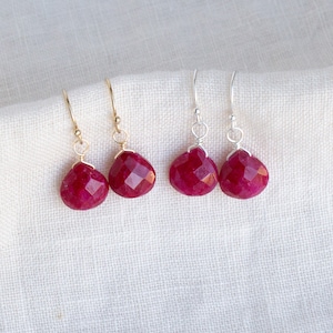 May include: Four pairs of dangle earrings with faceted, teardrop-shaped, deep red gemstones. Two pairs have gold-colored ear wires, and two pairs have silver-colored ear wires. The earrings are displayed on a white, textured fabric.