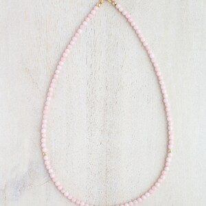 May include: A delicate pink beaded necklace with gold accents. The necklace features small, round pink beads strung together, with gold beads interspersed throughout. A gold-colored crescent moon charm is at the clasp.