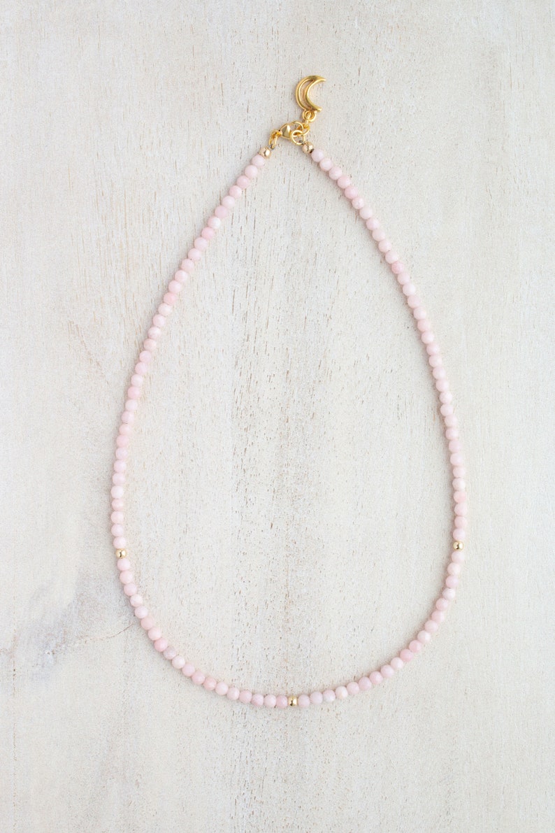 May include: A delicate pink beaded necklace with gold accents. The necklace features small, round pink beads strung together, with gold beads interspersed throughout. A gold-colored crescent moon charm is at the clasp.