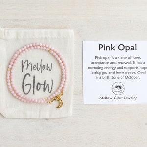 May include: A delicate pink opal necklace with a gold crescent moon charm, displayed in a cream-colored drawstring bag. The bag has the words "Mellow Glow" printed on it. A card next to it reads "Pink Opal".
