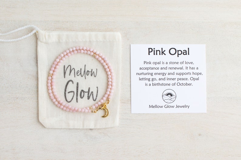 May include: A delicate pink opal necklace with a gold crescent moon charm, displayed in a cream-colored drawstring bag. The bag has the words "Mellow Glow" printed on it. A card next to it reads "Pink Opal".