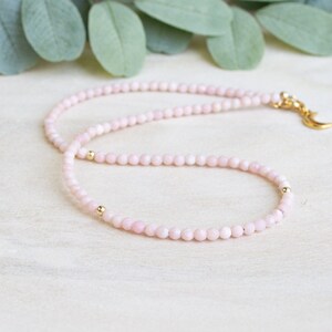 May include: A delicate pink beaded necklace with gold accents. The necklace features small, round pink beads and gold-colored beads. A gold-colored crescent moon charm is attached to the clasp. The necklace is displayed on a light-colored surface with green leaves in the background.